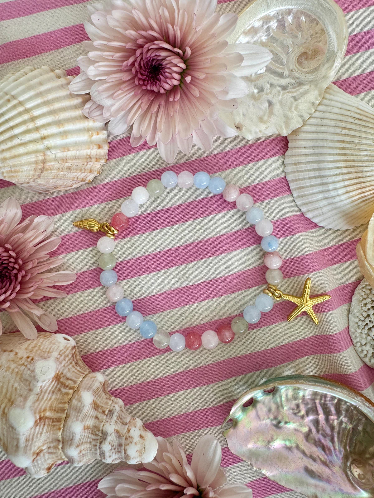 Handcrafted ocean-inspired Kiki Bracelet featuring pastel beads and gold starfish charm, surrounded by shells and flowers.