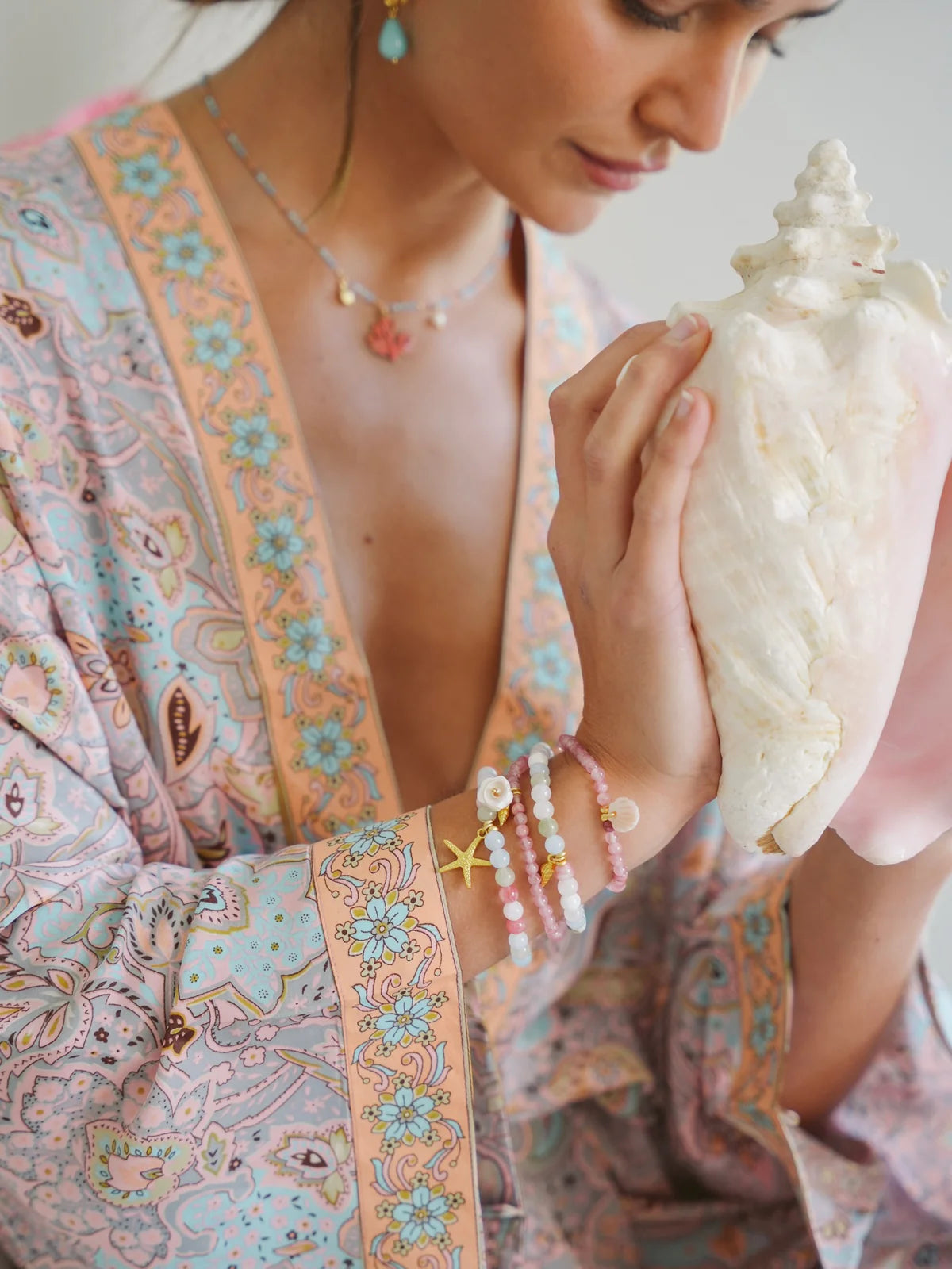 Woman holding a seashell, wearing handcrafted ocean inspired jewelry and a pastel patterned robe.